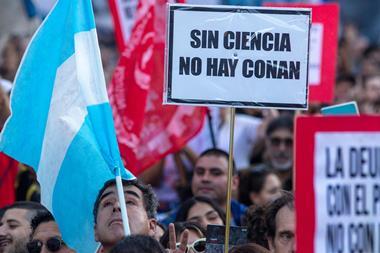 A large group of protesters. One is holding an Argentinian flag without the sun and one is holding a placard that says Sin Ciencia No Hay Conan (Without Science we have no Conan)